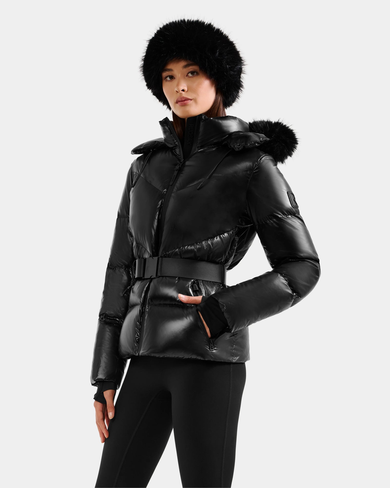 Wearing the Rudsak CHRISTELLE S - L125544 Black glossy down puffer with a belt, black leggings, and a faux fur hat, she stands against a light background—an effortlessly chic cold-weather look.