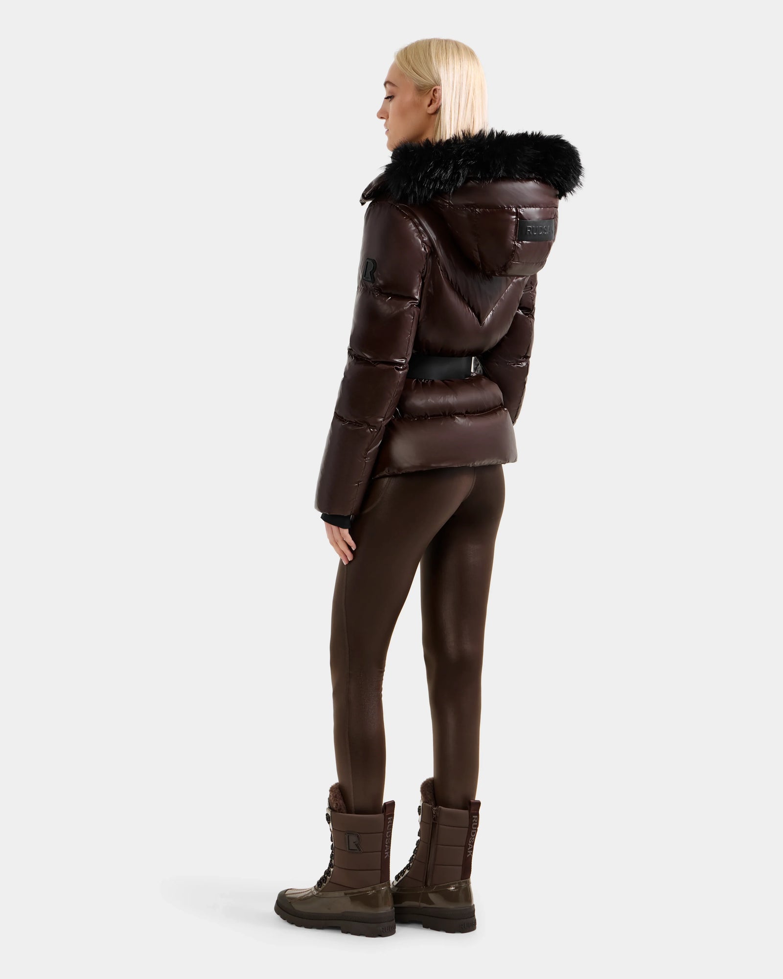 A person with straight blonde hair is facing away, wearing the Rudsak CHRISTELLE S - L125544 Espresso women’s down puffer with black faux fur hood. They pair it with matching leggings and boots against a plain light gray background.