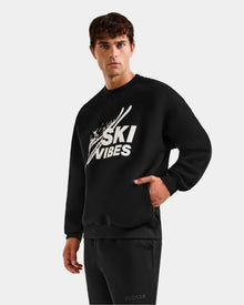 A young man in the Rudsak COLSON - M525501 Black men’s crewneck sweatshirt with SKI VIBES logo and ski graphics stands against a plain background, one hand in his pocket, exuding effortless après-ski style as he looks at the camera.