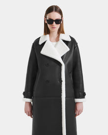 A person models the Rudsak DARCY - L125539 Black women's faux sherpa coat with white trim, standing against a plain white background and looking straight ahead with a neutral expression.