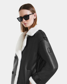 A woman in black sunglasses and the Rudsak DARCY - L125539 black double-breasted winter coat with a white shearling collar stands against a plain, light background.