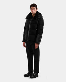 A man stands against a plain light background wearing the Rudsak DAVID - M125508 Black men’s shearling down puffer with leather sleeves, black pants, and black boots, facing slightly left with a neutral expression, showcasing winter-ready style.