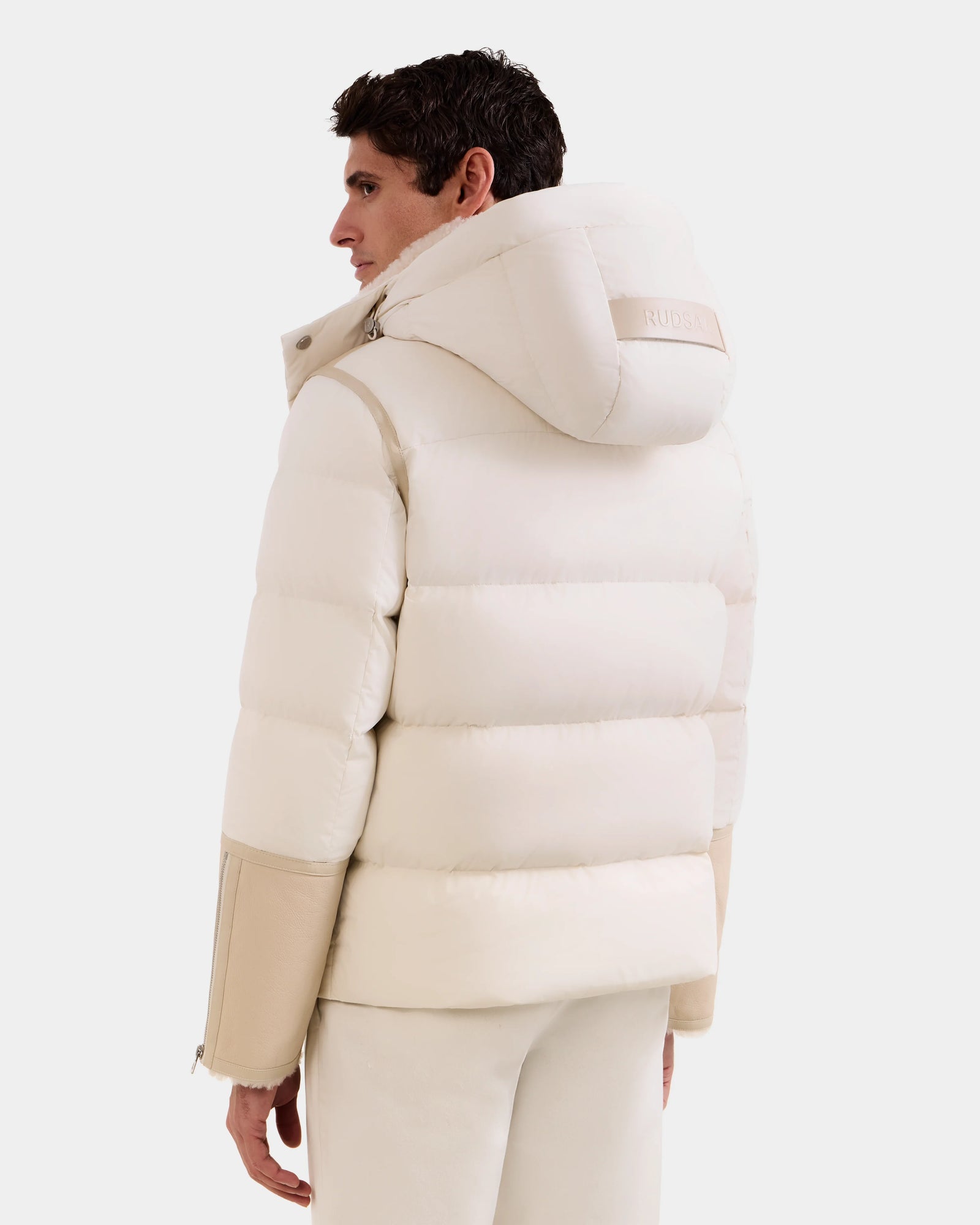 A person is seen from the back, looking to the side against a white background, wearing the Rudsak DAVID - M125508 Vanilla puffy lamb shearling hooded winter jacket with beige sleeves and white pants.