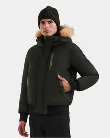 A man wearing the Rudsak DEACON - M123513 Dark Olive bomber-style down puffer with a fur-lined hood, paired with a black knit beanie and black pants, stands with one hand in his pocket against a plain light background.
