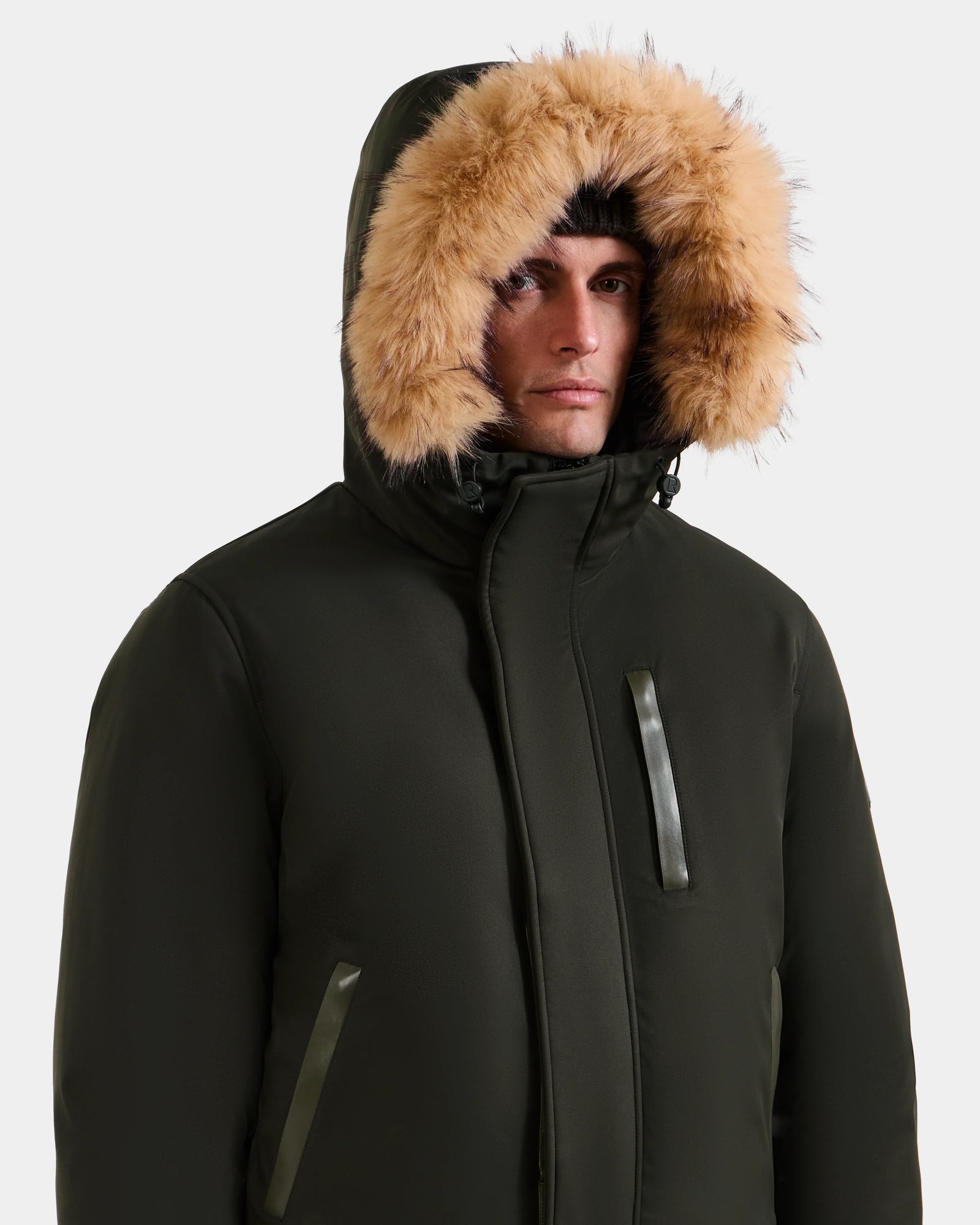 A person wears the Rudsak DEACON - M123513 Dark Olive bomber-style down puffer with a fur-lined hood, fully zipped, and looks slightly to the side against a plain light background.