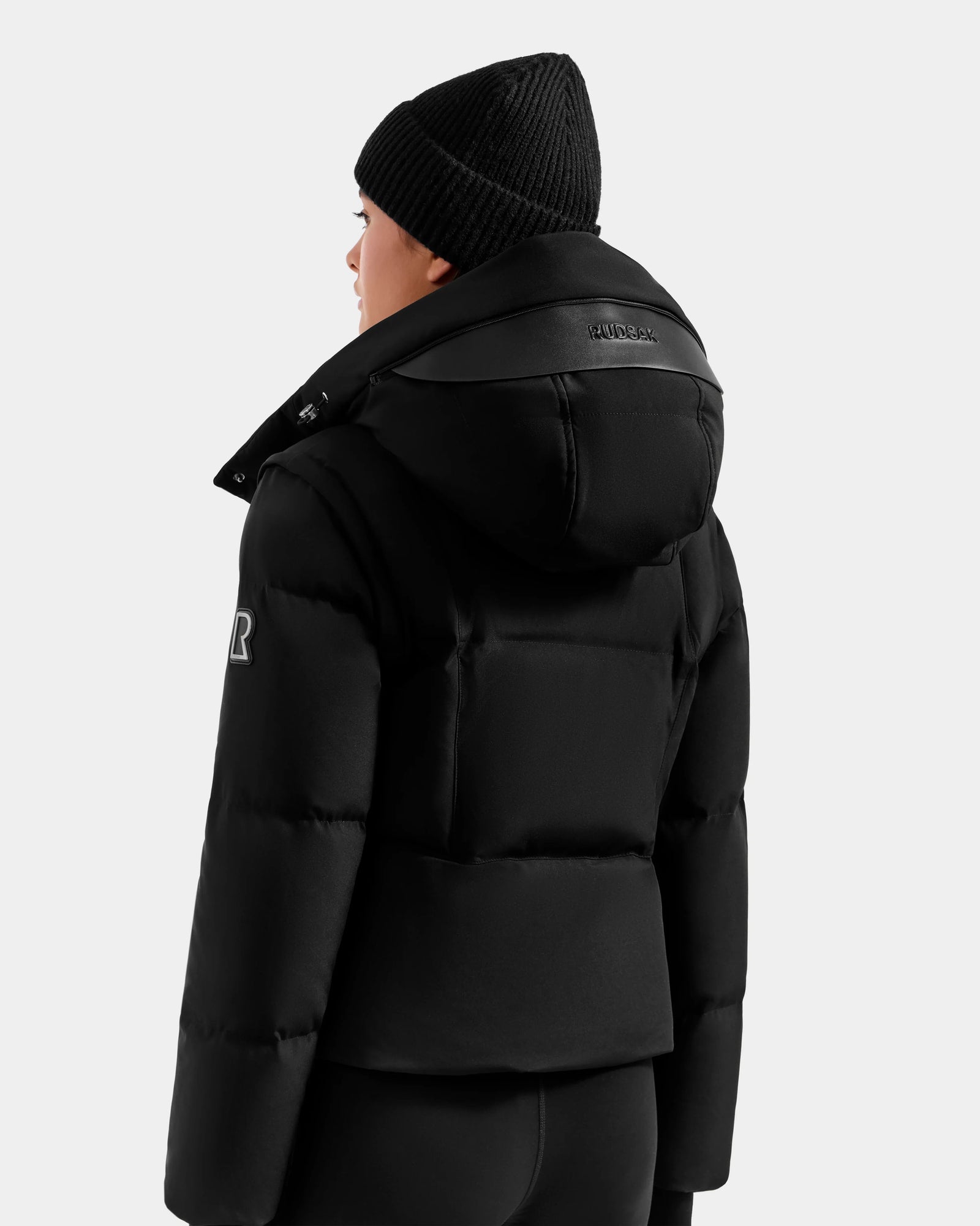 A person is seen from the back and side against a plain light background wearing Rudsak’s DELILAH M - L125538 Black women’s down puffer jacket with hood, visible logo on the sleeve and collar, black knit beanie, and black pants.