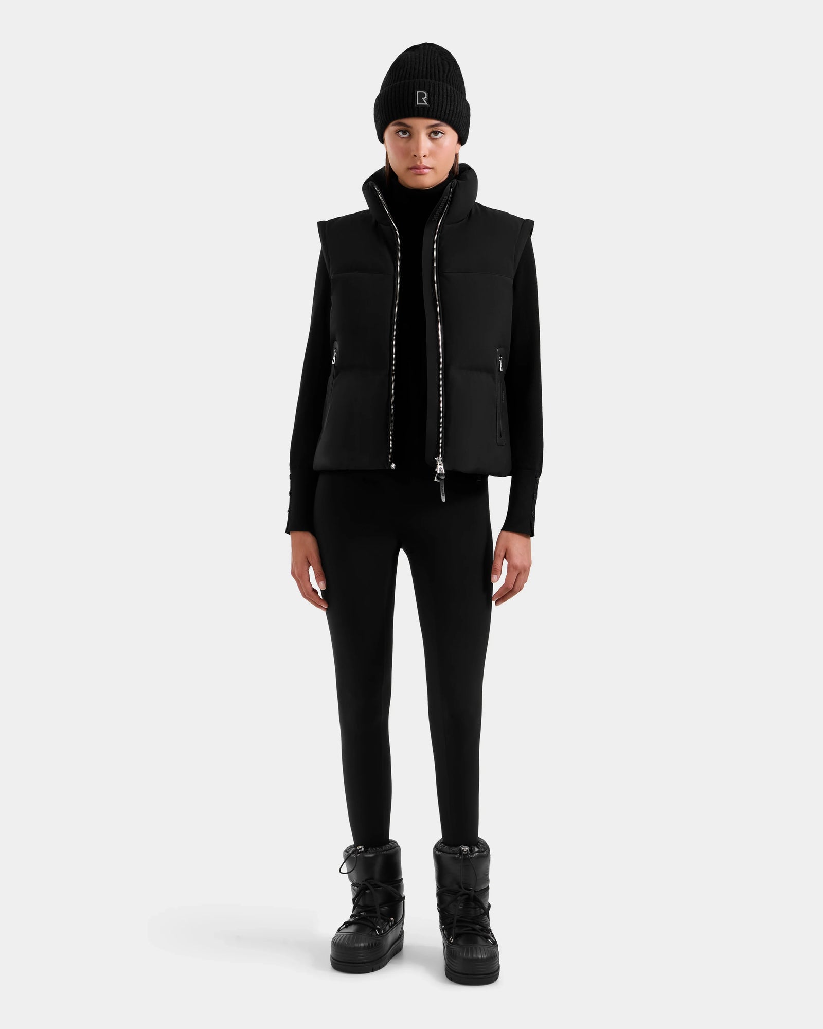 A person stands facing forward wearing the Rudsak DELILAH M - L125538 Black women’s down puffer with removable sleeves, a black long-sleeve top, leggings, and lace-up boots against a plain light gray background.