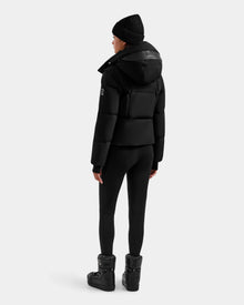 A person wearing a Rudsak DELILAH M - L125538 Black women’s down puffer with removable sleeves, black leggings, winter boots, and a black beanie stands facing away from the camera against a plain light gray background.