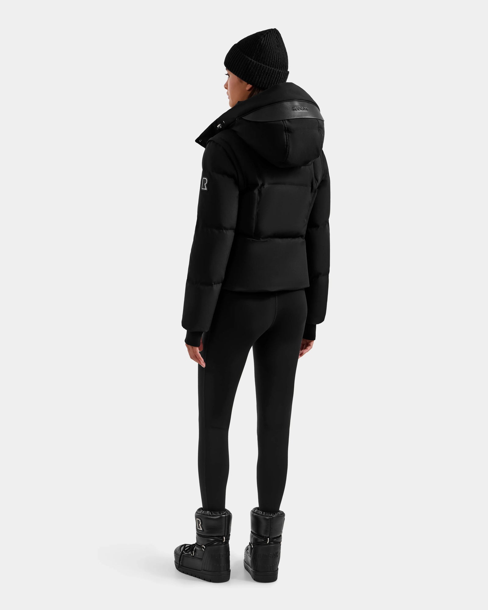 A person wearing a Rudsak DELILAH M - L125538 Black women’s down puffer with removable sleeves, black leggings, winter boots, and a black beanie stands facing away from the camera against a plain light gray background.