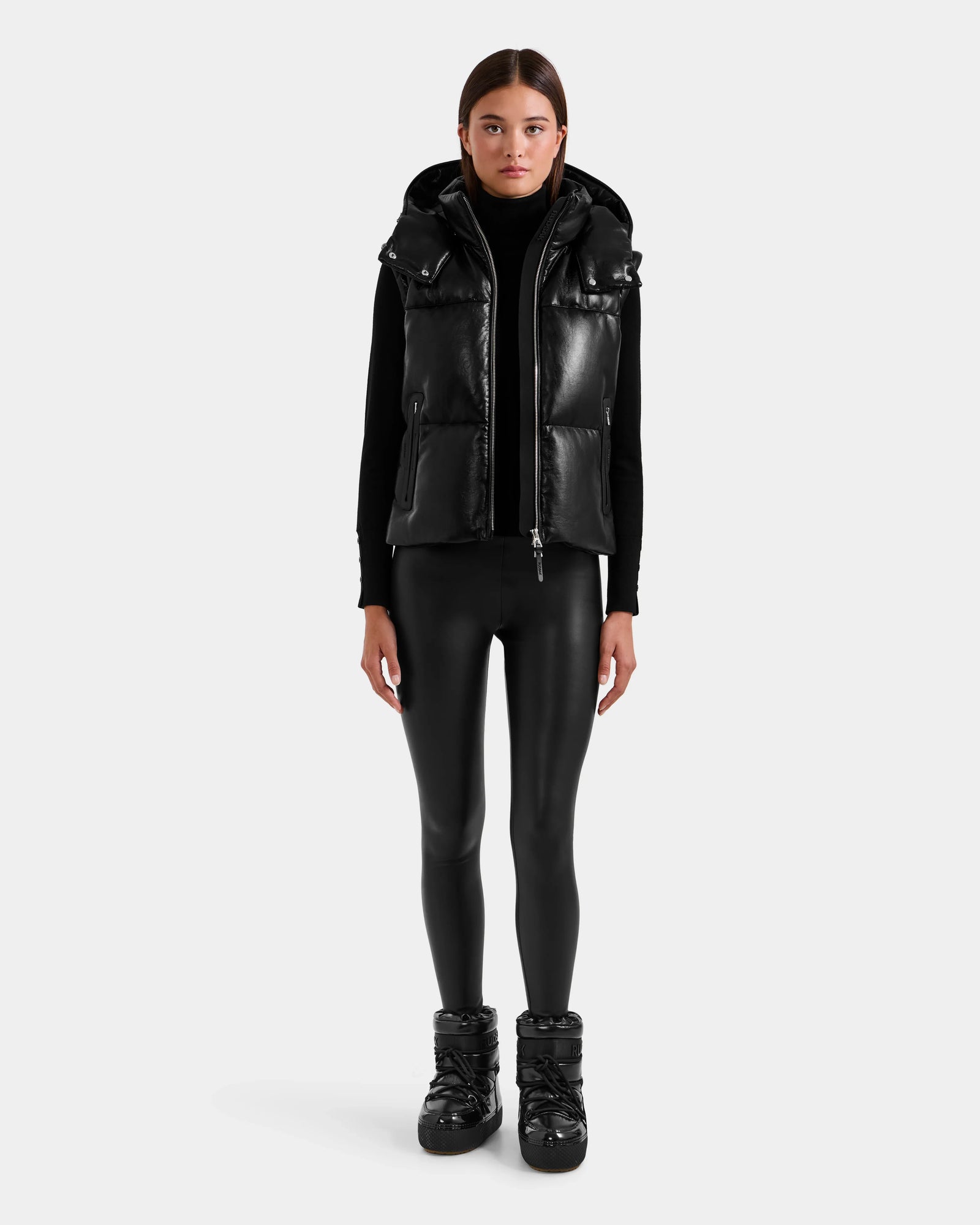 A person stands facing forward in the Rudsak DELILAH PX - L125537 Black women’s vegan-leather puffer vest, paired with a black turtleneck, shiny black leggings, and black boots against a plain white background.