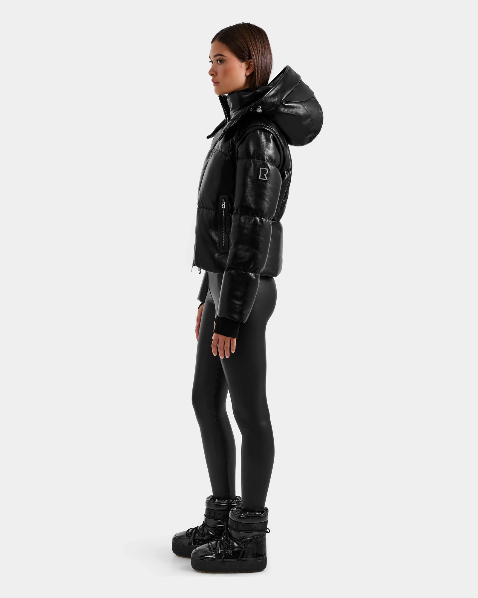 A woman stands in profile wearing the Rudsak DELILAH PX - L125537 Black vegan-leather puffer jacket with a large hood, styled with black leggings and matching puffer boots against a plain light gray background.
