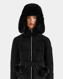A person wears the Rudsak DEMI - L425539 Black puffer coat with a belted waist and a Rudsak faux-fur aviator hat with ear flaps and pom-pom ties, standing against a plain light gray background.