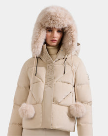 A woman in the Rudsak DEMI - L425539 Vanilla puffer jacket with a plush fur-trimmed hood and pom-pom drawstrings stands against a plain light background, channeling the cozy style of a women’s faux-fur aviator hat.