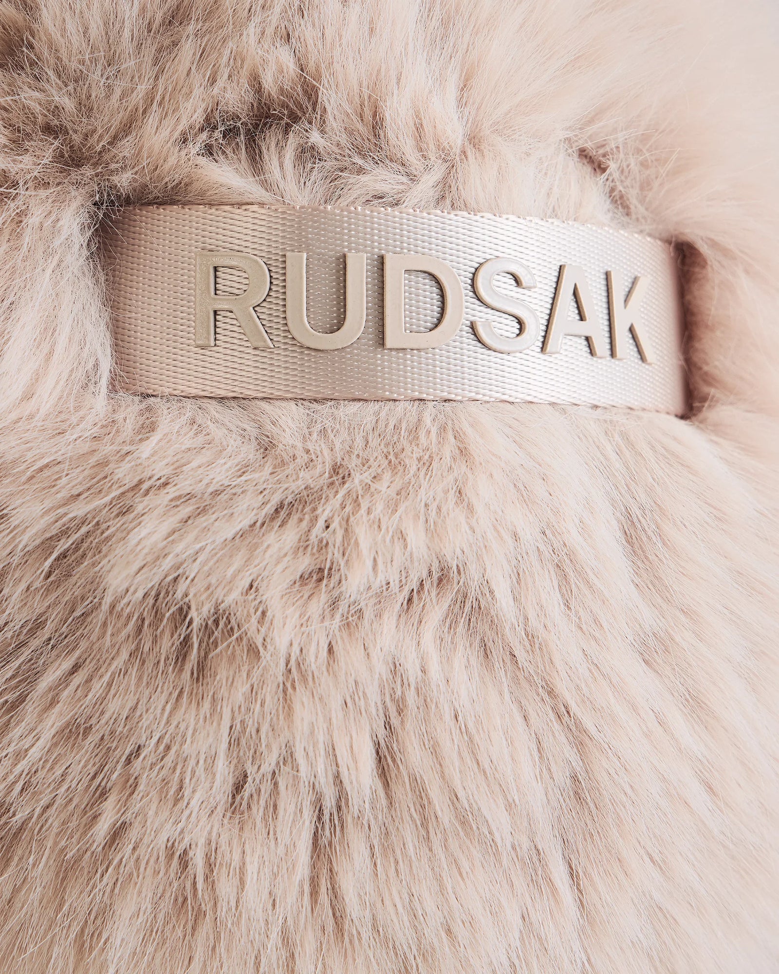 Close-up of soft, light beige faux fur on the DEMI - L425539 Vanilla women’s winter hat by Rudsak, featuring a shiny beige ribbon with “RUDSAK” in raised metallic letters on this luxe aviator style.