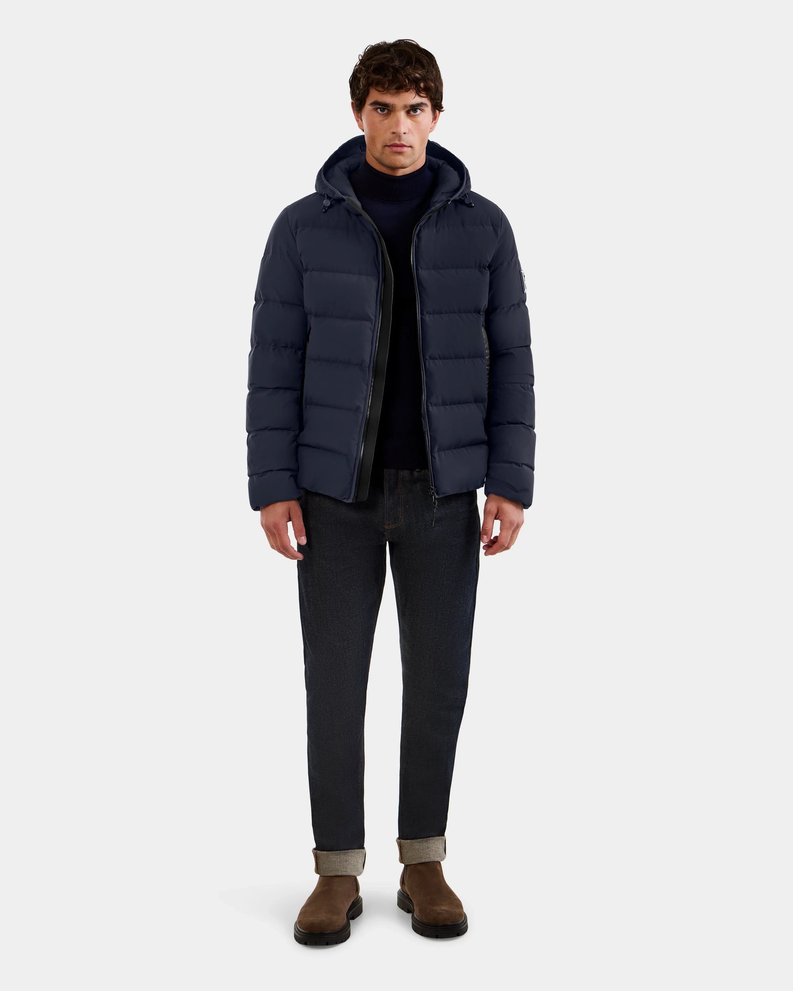 A man faces forward wearing the Rudsak DEMIAN M - M123507 Navy down puffer jacket, crafted from sustainable recycled fabric, styled with a dark sweater, cuffed black jeans, and brown suede boots against a light gray background.