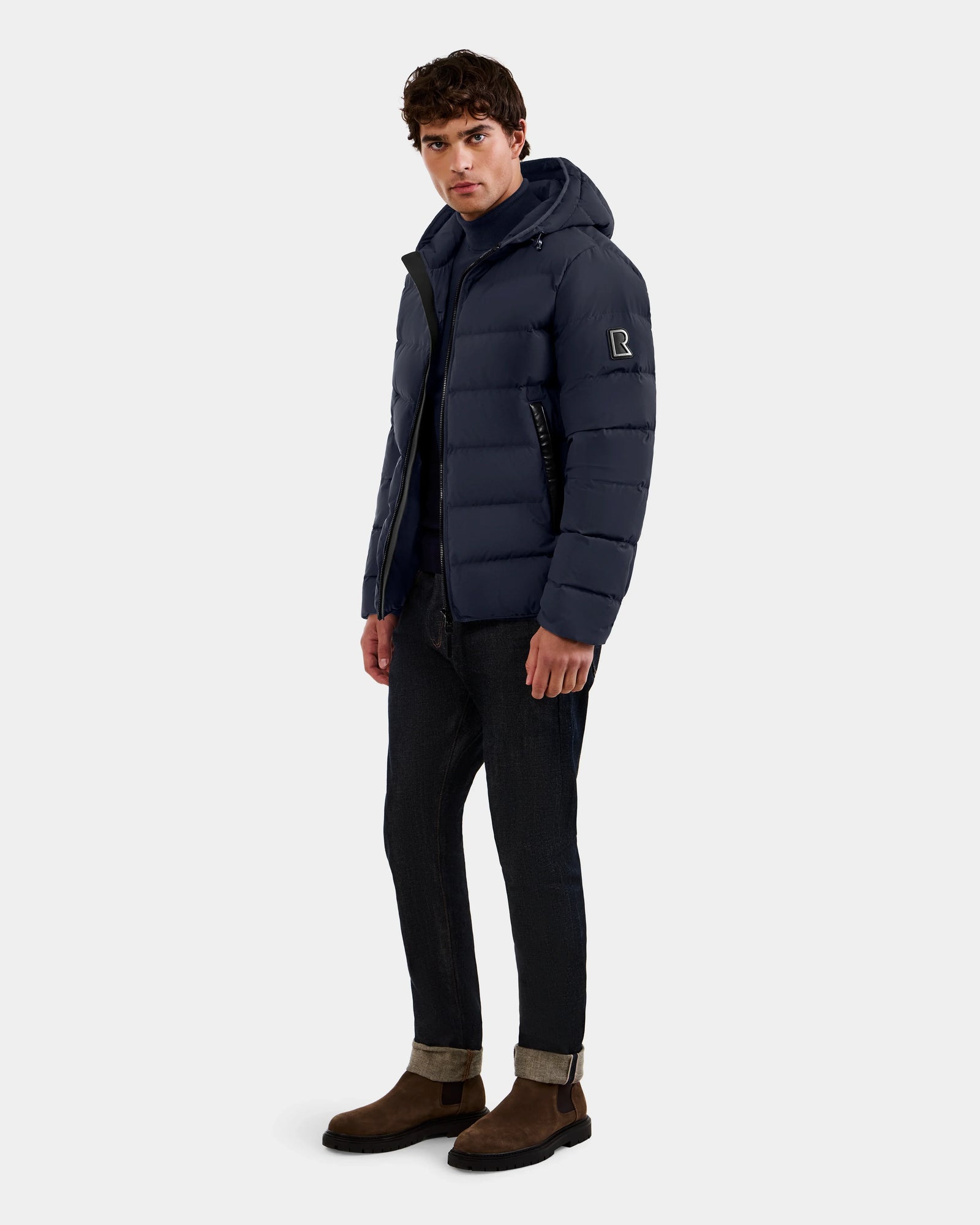 A man stands facing forward in the Rudsak DEMIAN M - M123507 Navy men’s down puffer jacket, made from RDS-certified premium down and recycled fabric. He pairs it with dark cuffed jeans and brown suede boots against a plain light gray background.