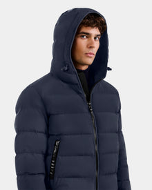 A young man wears the Rudsak DEMIAN M - M123507 Navy down puffer jacket with hood, featuring black zippers. Made from sustainable recycled fabric, it offers warmth and comfort against a light plain background.