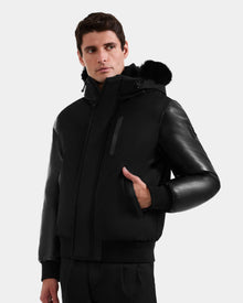 A man stands against a plain white background wearing the Rudsak DERON - M125539 Black down bomber with vegan-leather sleeves and a fur-trimmed hood, looking slightly to the side with one hand in his pocket.
