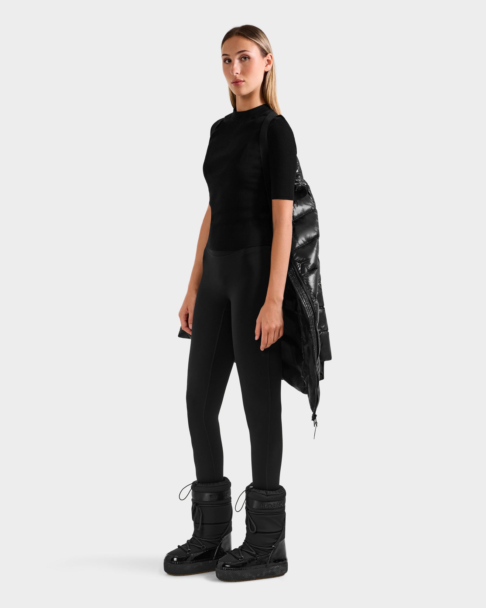 A woman wearing Rudsak’s ELISA S - L125572 Black stands against a plain background in black leggings, a fitted short-sleeve top, and winter boots. She drapes a mid-length coat over her arm and looks to the side with a neutral expression.