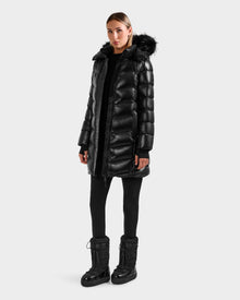 A woman stands against a plain background wearing the Rudsak ELISA S - L125572 Black quilted puffer jacket with a fur-trimmed hood, paired with black leggings and winter boots, looking straight ahead with a neutral expression.
