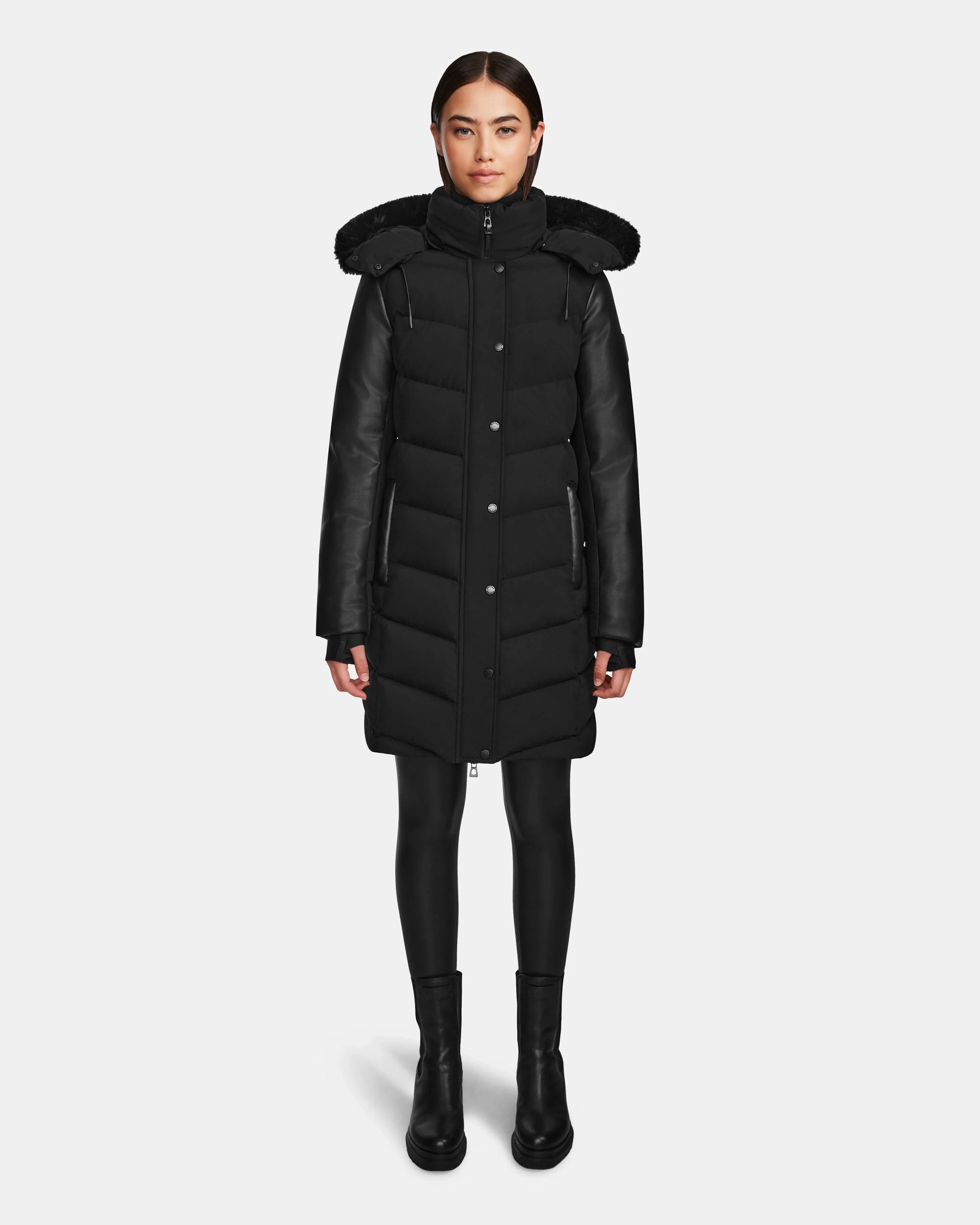 A woman faces forward against a white background, wearing the Rudsak ELSA - L123535 Black quilted down puffer winter coat with a hood, paired with black leggings and boots.