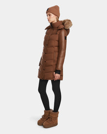 A woman in profile wears the Rudsak ELSA - L123535 Taupe: a quilted down puffer with a fur-trimmed hood, styled with a brown beanie, black leggings, and brown winter boots against a plain light background.