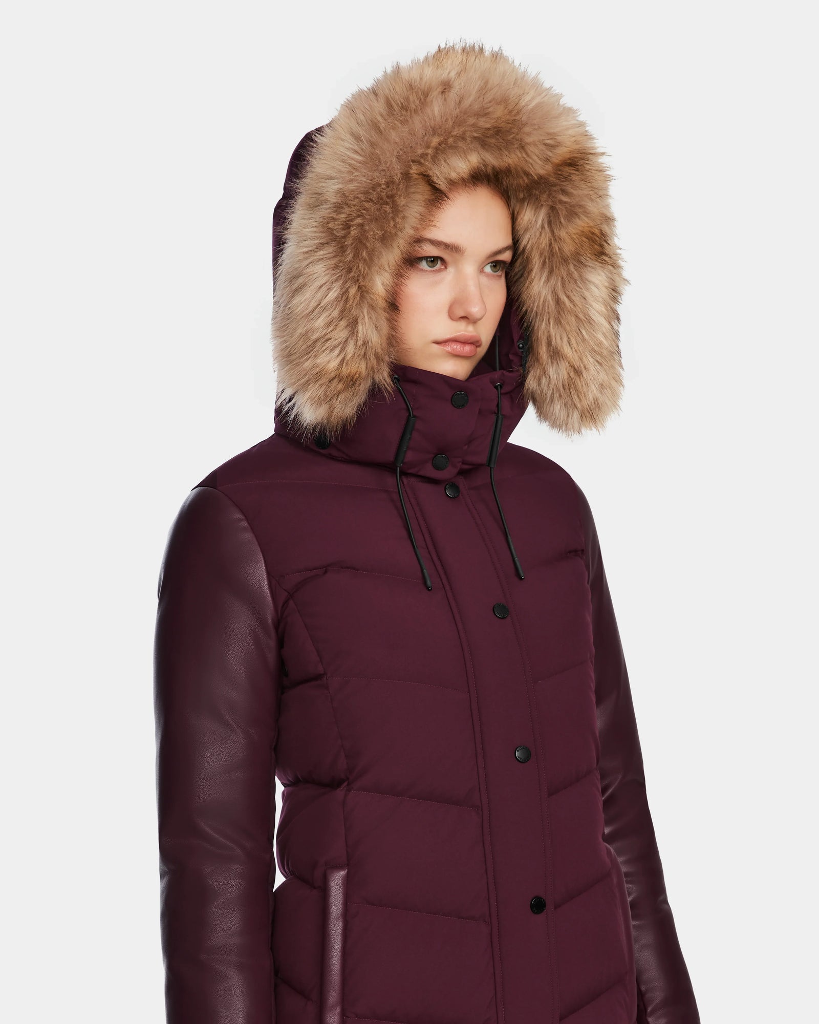 A person wears the Rudsak ELSA - L123535 Wine womens quilted down puffer with leather sleeves and a large faux fur-trimmed hood, standing against a plain light background.