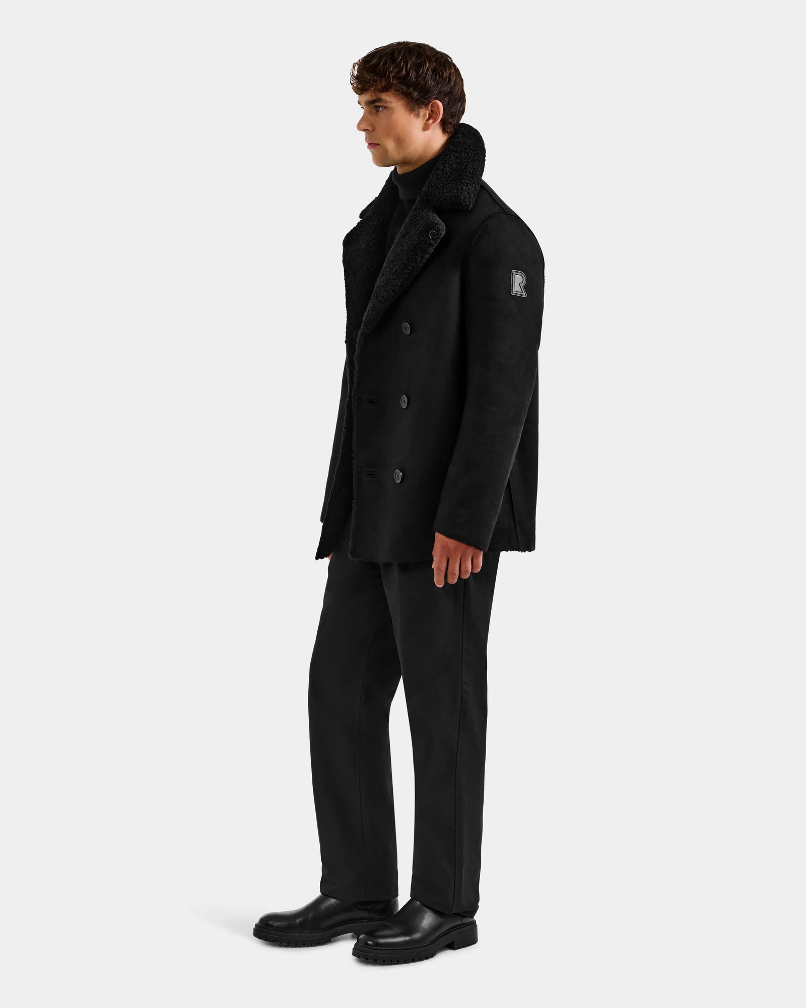 A man stands in profile against a plain light background, wearing the Rudsak ELTON - M125534 Black double-breasted overcoat with faux shearling, paired with a black turtleneck, pants, and shoes.
