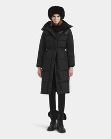 A person stands facing forward wearing the Rudsak EMBER M-L - L125543 Black women’s long down puffer coat with a removable hood, paired with black pants, lace-up fur-trimmed boots, a furry hat, and a scarf against a plain white background.