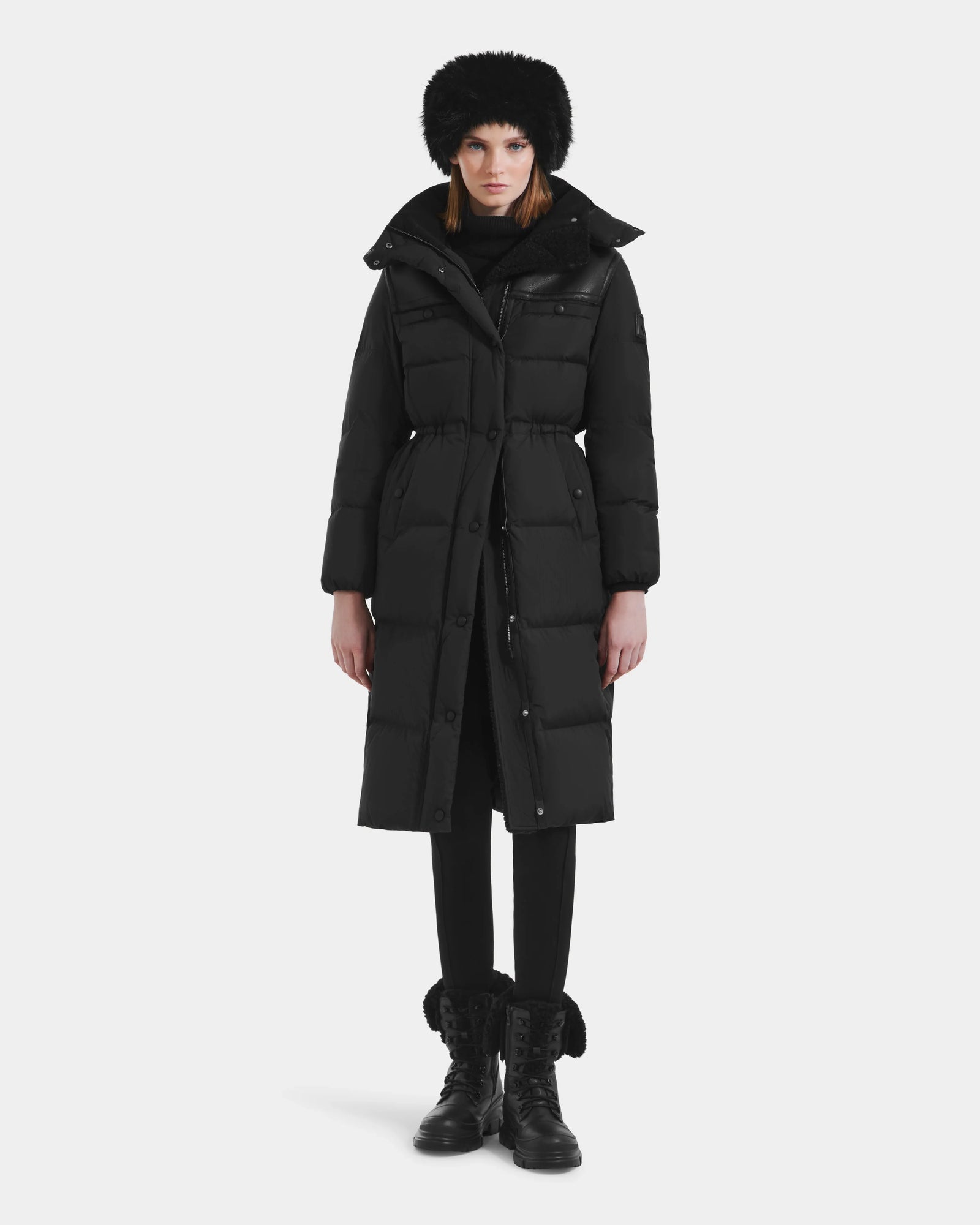 A person stands facing forward wearing the Rudsak EMBER M-L - L125543 Black women’s long down puffer coat with a removable hood, paired with black pants, lace-up fur-trimmed boots, a furry hat, and a scarf against a plain white background.