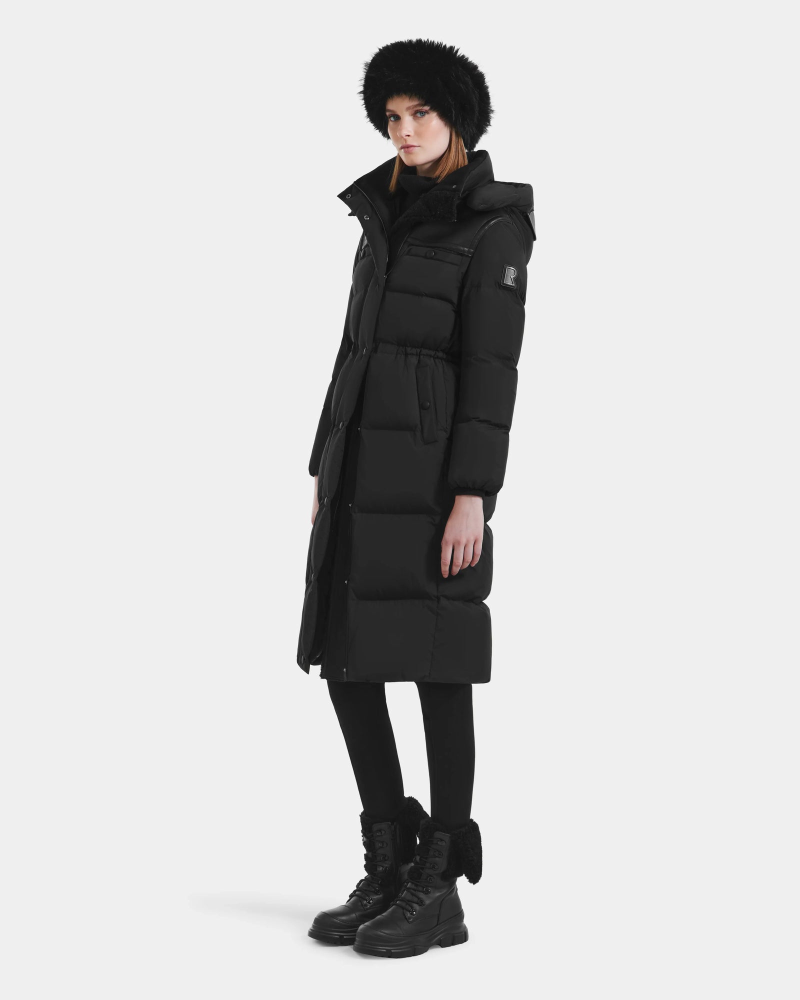 A person wearing the Rudsak EMBER M-L - L125543 Black women’s long down puffer with removable hood, a black furry hat, black leggings, and black boots stands against a plain white background.