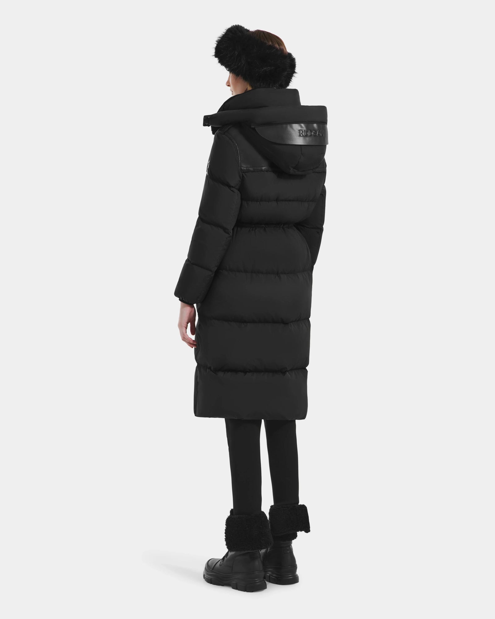 A person stands facing away, wearing the Rudsak EMBER M-L - L125543 Black women’s long down puffer coat with removable hood, paired with black leggings, black furry boots, and a black fur headband against a plain light background.