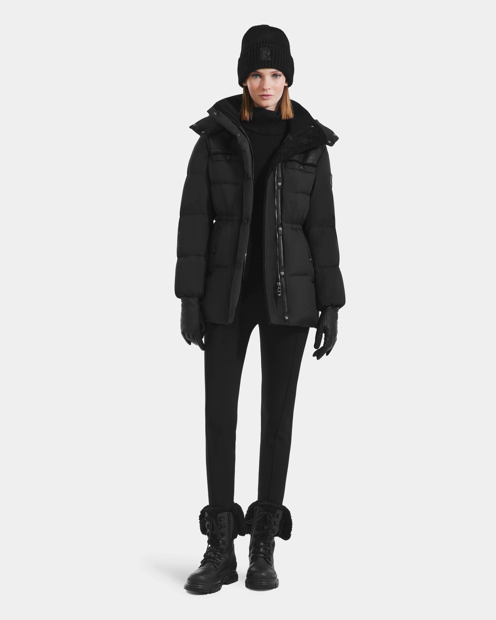 A person stands facing forward in the Rudsak EMBER M-M - L125524 Black women’s down puffer with vegan-leather piping, paired with a black beanie, gloves, pants, and fur-trimmed lace-up boots against a plain white background.