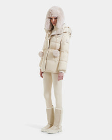 A woman stands against a white background wearing the Rudsak EMBER M-M - L125524 down puffer in Vanilla, featuring RDS-certified down, fur trim, a matching hat, slim pants, and tall beige boots.