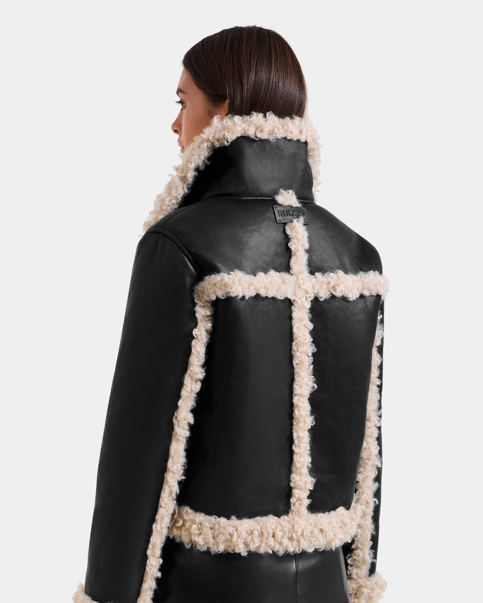 A woman with straight brown hair is seen from the back wearing the Rudsak EMERY - L125548 Black vegan-leather jacket, featuring a high collar, white faux shearling trim, and decorative seams outlined in soft shearling.