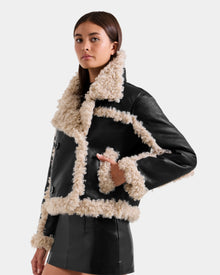A woman models the Rudsak EMERY - L125548 Black vegan-leather jacket with cream faux shearling trim and a matching black mini skirt, standing against a plain light background.