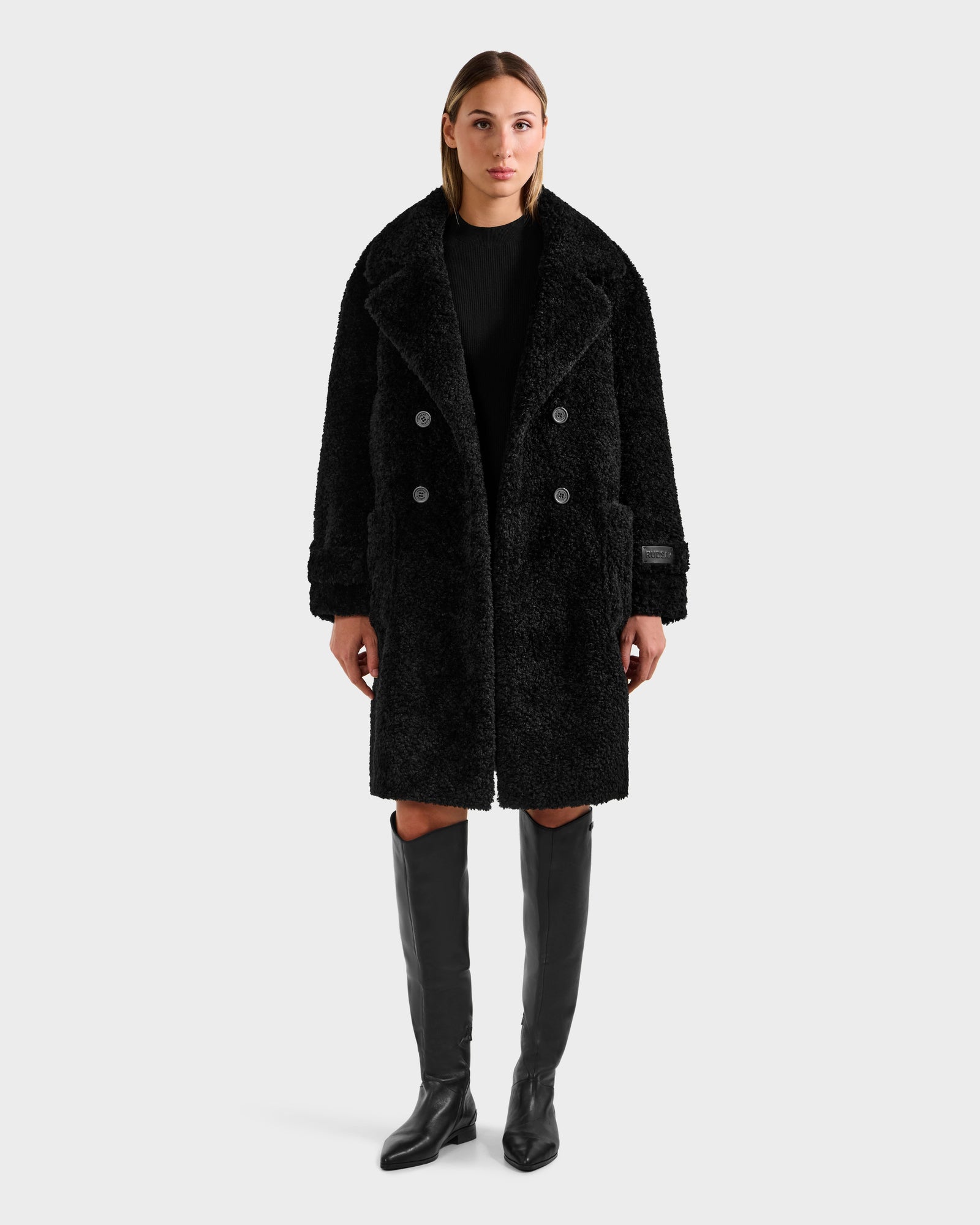 A woman faces forward wearing the Rudsak EMRYN - L125588 Black textured winter coat, styled like a teddy jacket, with black knee-high boots and a black top against a light gray background.