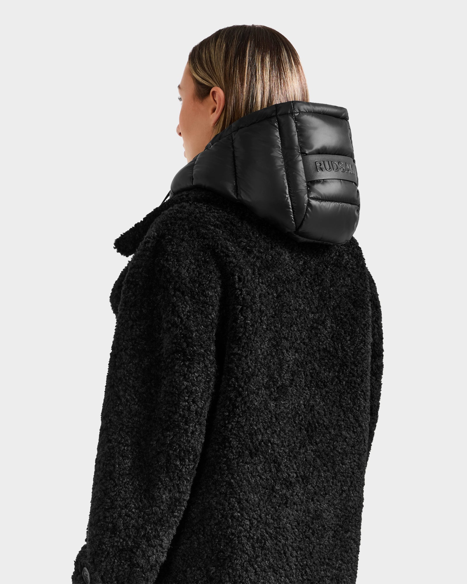 A person with straight, light brown hair is shown from behind wearing the Rudsak EMRYN - L125588 Black winter coat, featuring a large shiny hood embossed with RUDSAK, echoing the cozy look of a women’s teddy jacket against a light background.