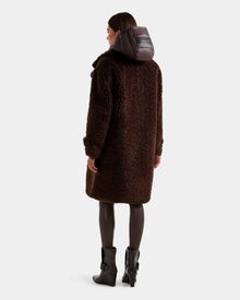 A woman faces away, wearing the Rudsak EMRYN - L125588 Espresso long brown teddy jacket with a dark hood, black leggings, and heeled black ankle boots against a plain light background.