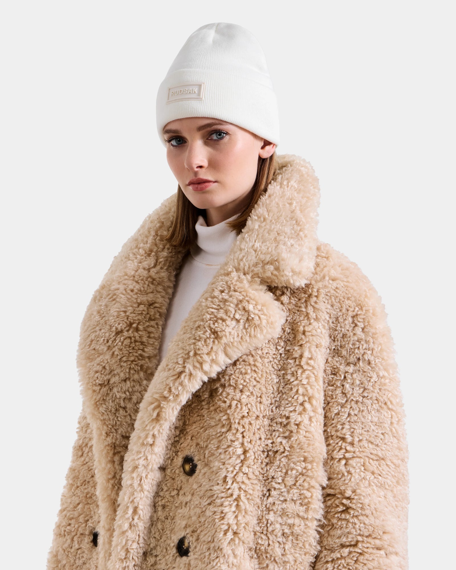 A woman stands against a plain light background wearing the Rudsak EMRYN - L125588 Vanilla teddy jacket with large lapels and black buttons, paired with a white beanie.