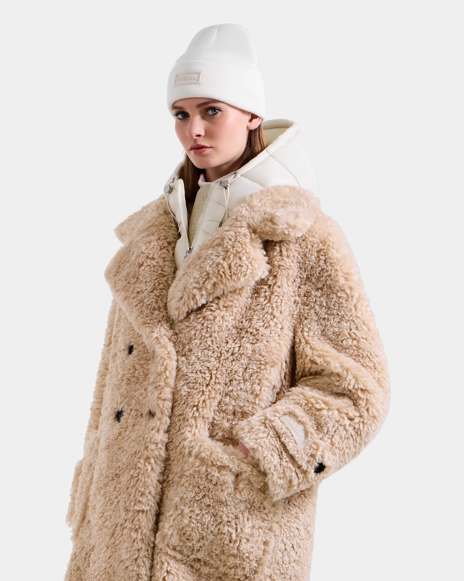 A woman models the Rudsak EMRYN - L125588 Vanilla, a large beige winter teddy coat, over a white puffer jacket and beanie. She stands against a plain light grey background, gazing slightly to the side.