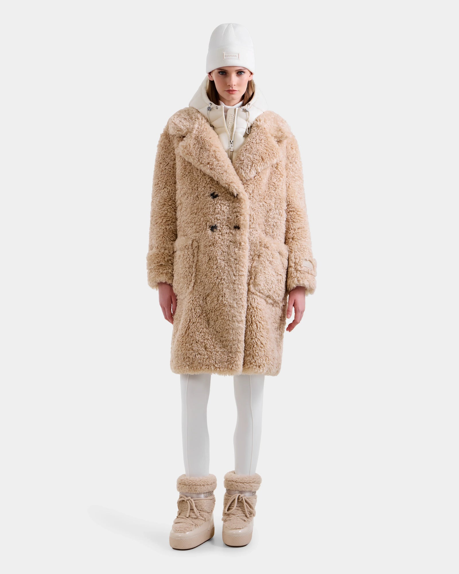 A person stands facing forward in a Rudsak EMRYN - L125588 Vanilla women’s teddy jacket, styled with white leggings, beige fur boots, a white hoodie, and a white cap against a plain white background.