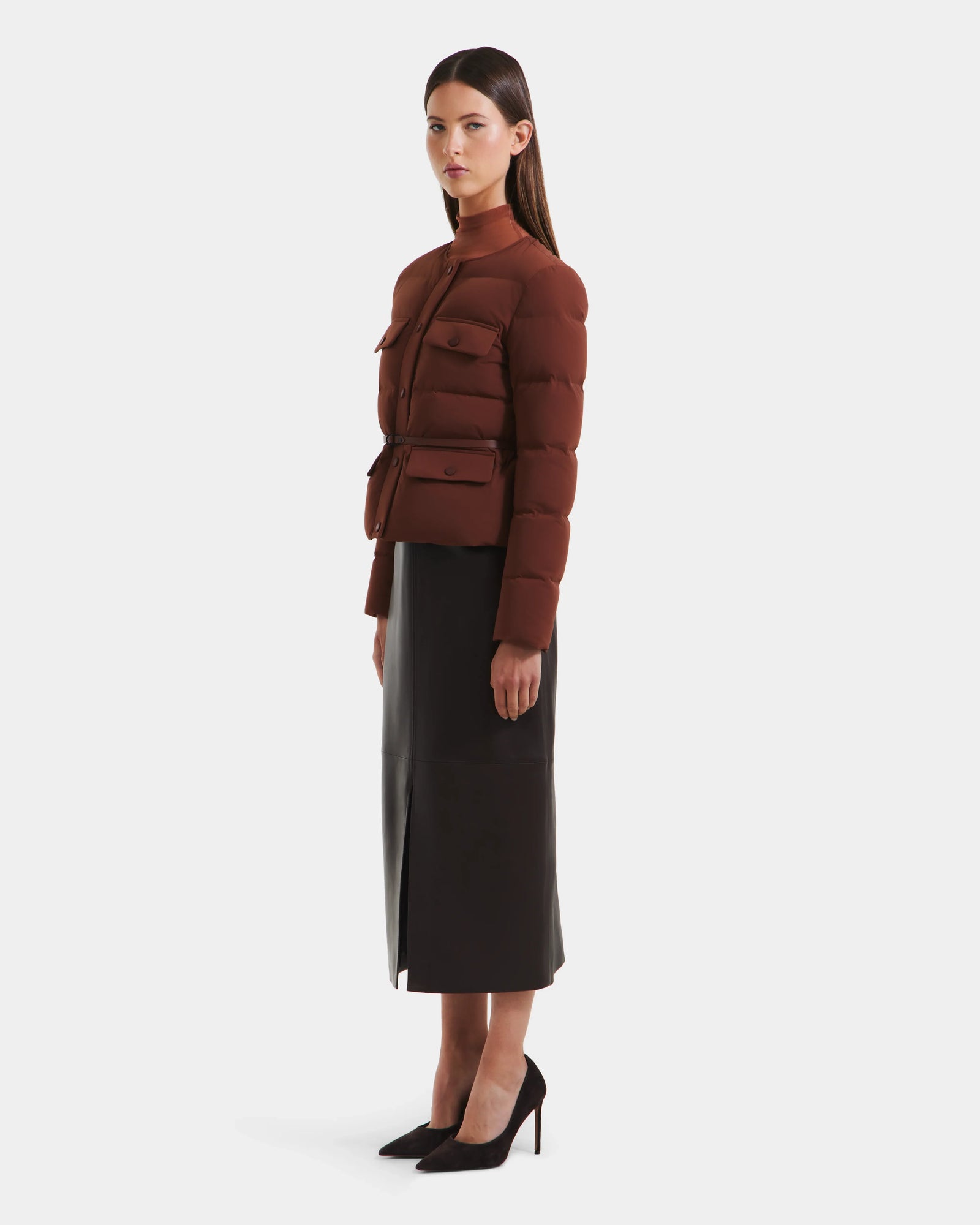 A woman stands sideways in a Rudsak FRANCESCA - 8125021 Rust fitted, down-quilted puffer jacket with a vegan leather belt, styled with a black midi skirt and high heels, against a plain white background.