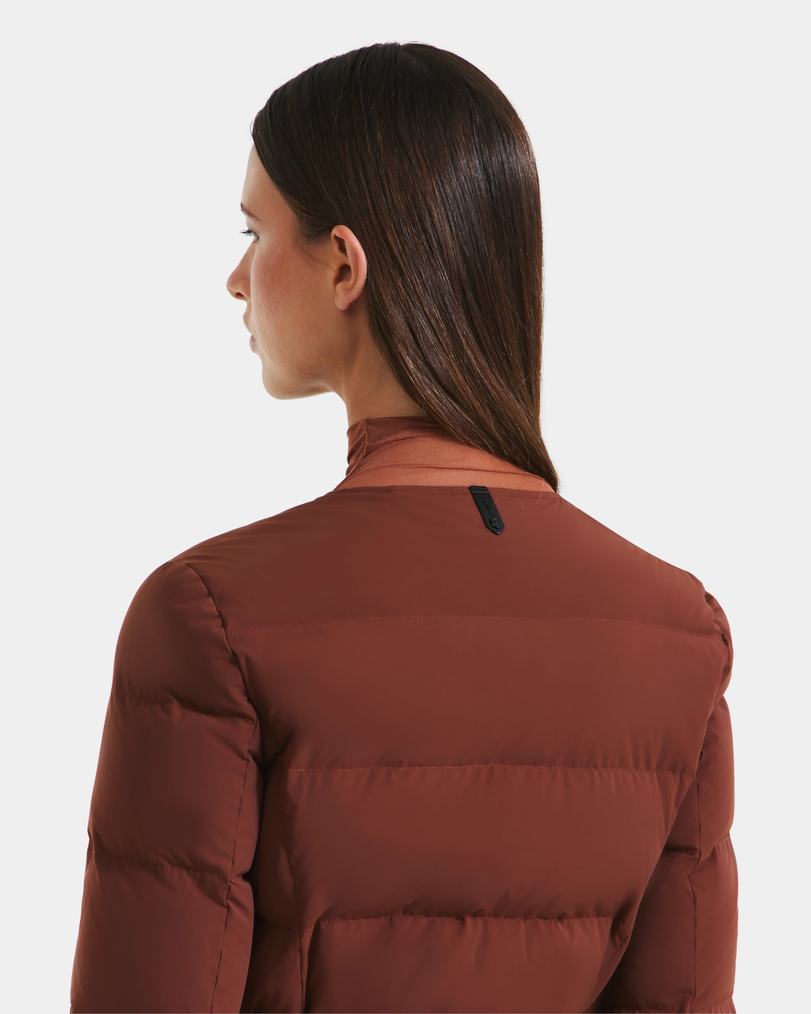 A woman with straight brown hair models the Rudsak FRANCESCA - 8125021 Rust quilted puffer jacket, facing away to reveal the back and side view against a plain white background.