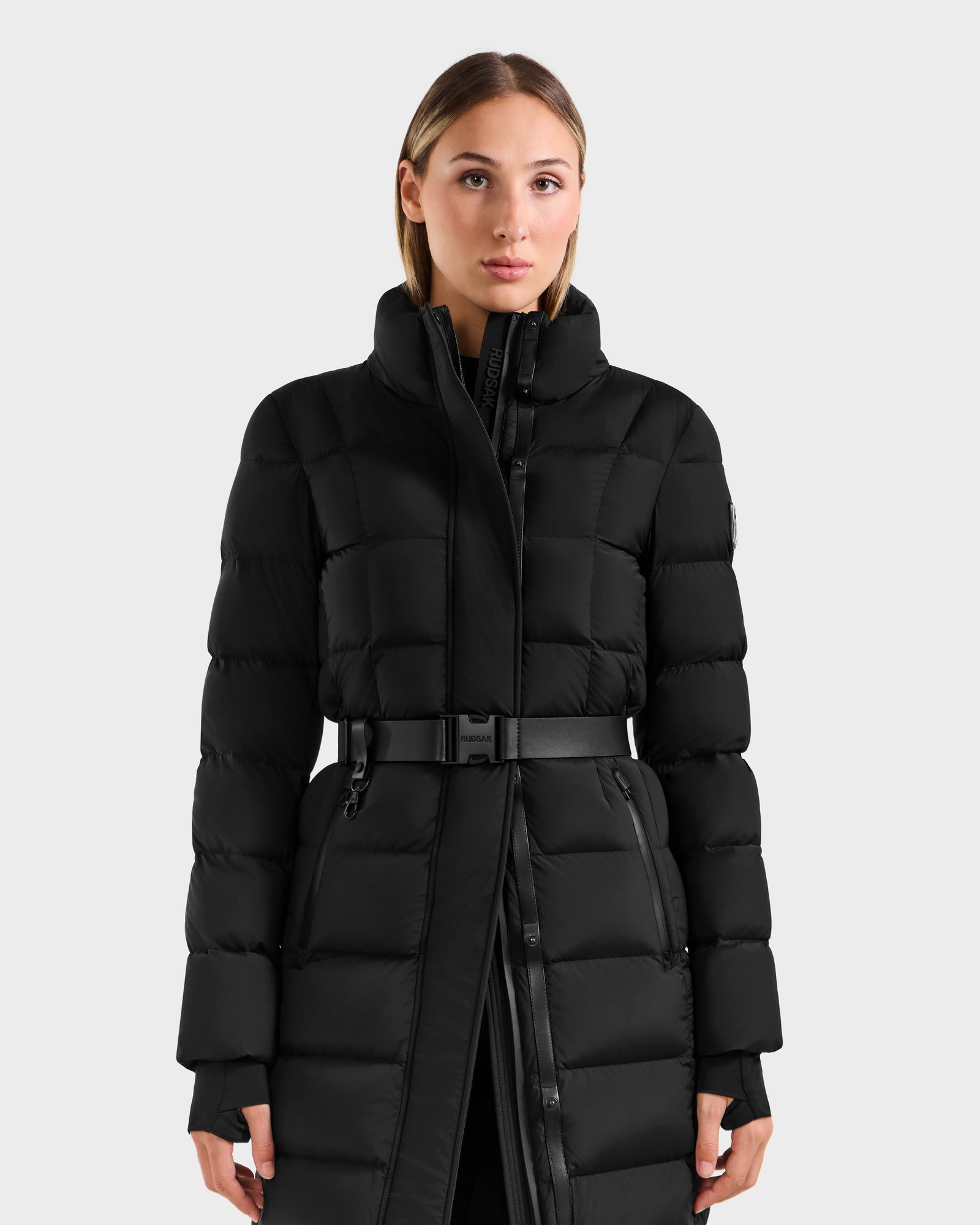 A woman with straight, light brown hair models the Rudsak FRIDA M-L - L125608 Black women’s long down puffer coat, featuring a high collar and belted waist, against a plain light gray background.