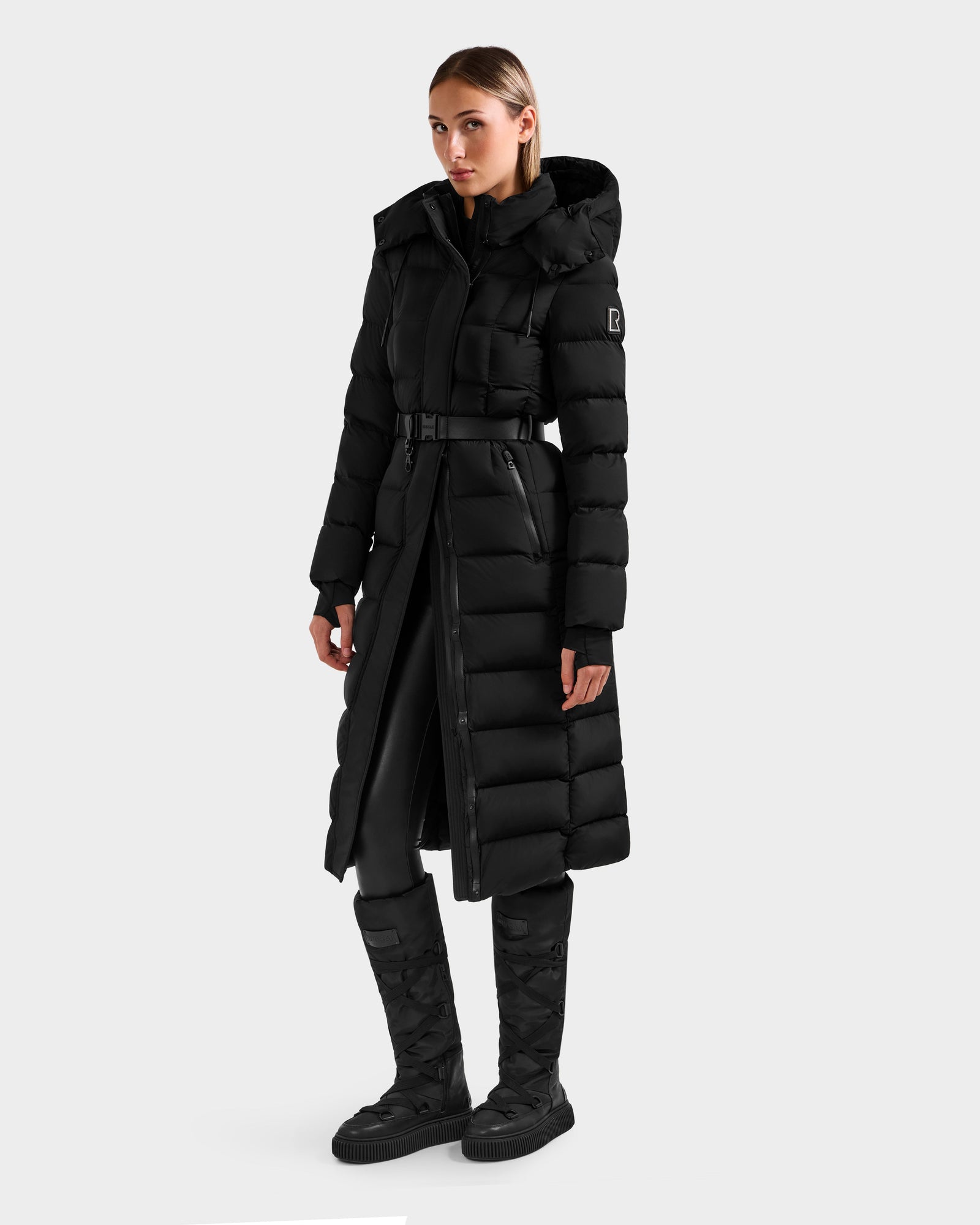 A woman in the Rudsak FRIDA M-L - L125608 Black long down puffer with detachable hood, paired with black pants and tall black quilted boots, stands against a plain light-gray background.