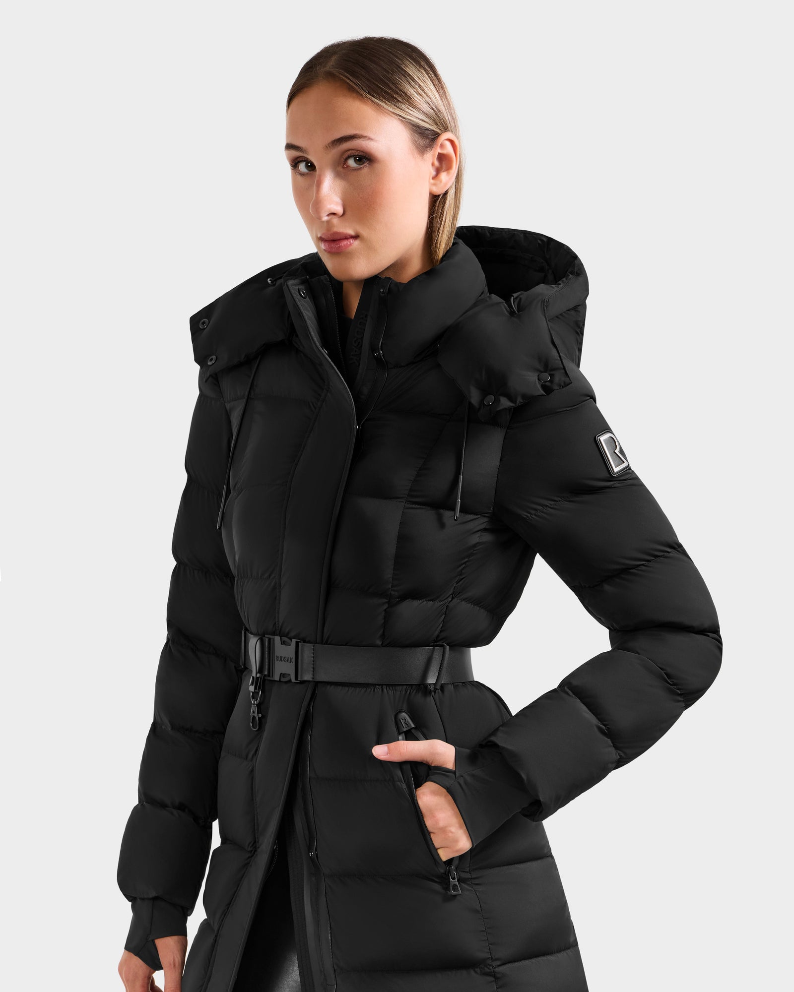 A person stands facing forward, one hand in a pocket, wearing the Rudsak FRIDA M-L - L125608 Black belted women’s long down puffer with hood against a plain light background.