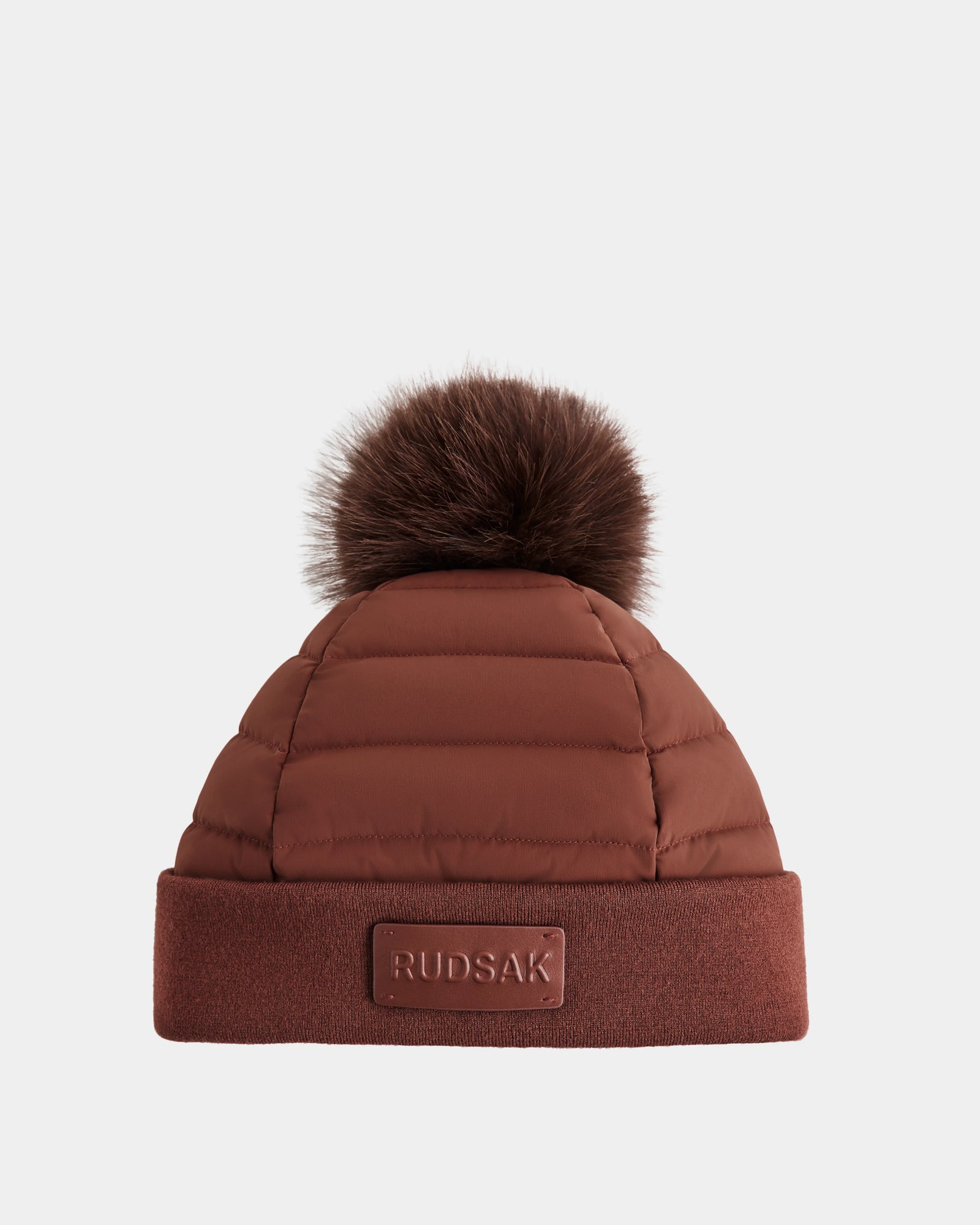 The Rudsak GALA - 8424514 Rust is a women's brown quilted down puffer hat with a large removable faux fur pom pom and folded brim featuring a rectangular RUDSAK patch.