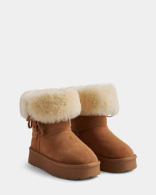 The Rudsak INESA - L225026 Tan boots feature brown suede, a fluffy white wool lining at the top, side bow detail, and flat rubber soles, shown against a plain light gray background.