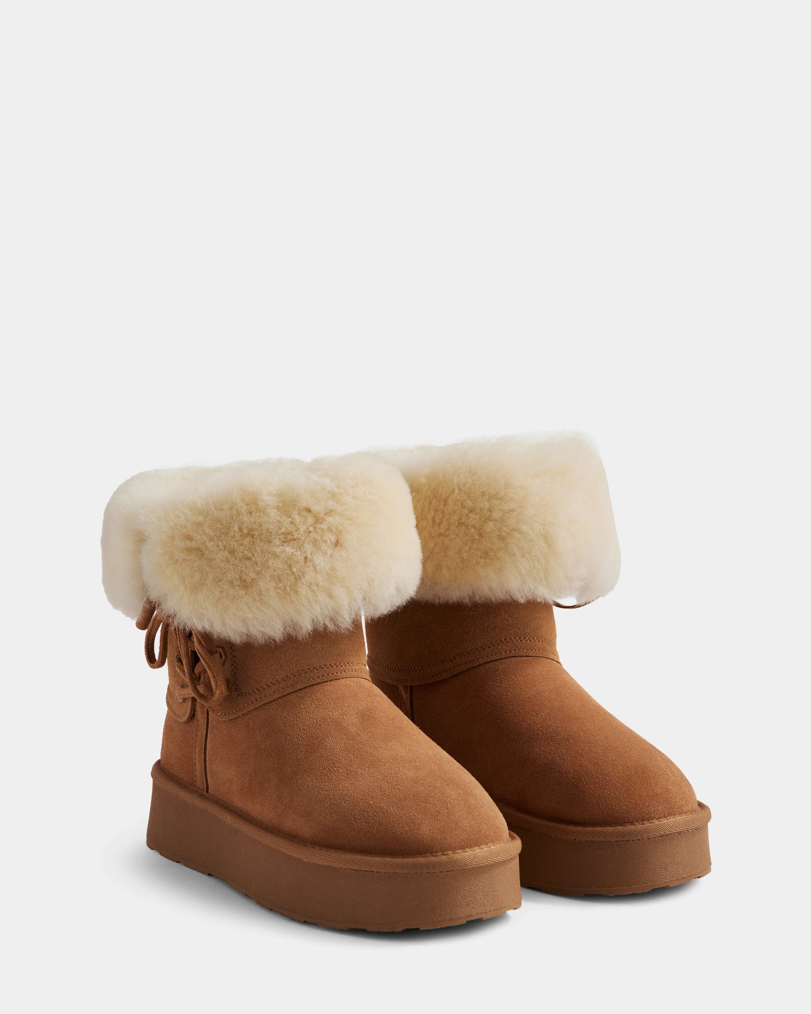 The Rudsak INESA - L225026 Tan boots feature brown suede, a fluffy white wool lining at the top, side bow detail, and flat rubber soles, shown against a plain light gray background.